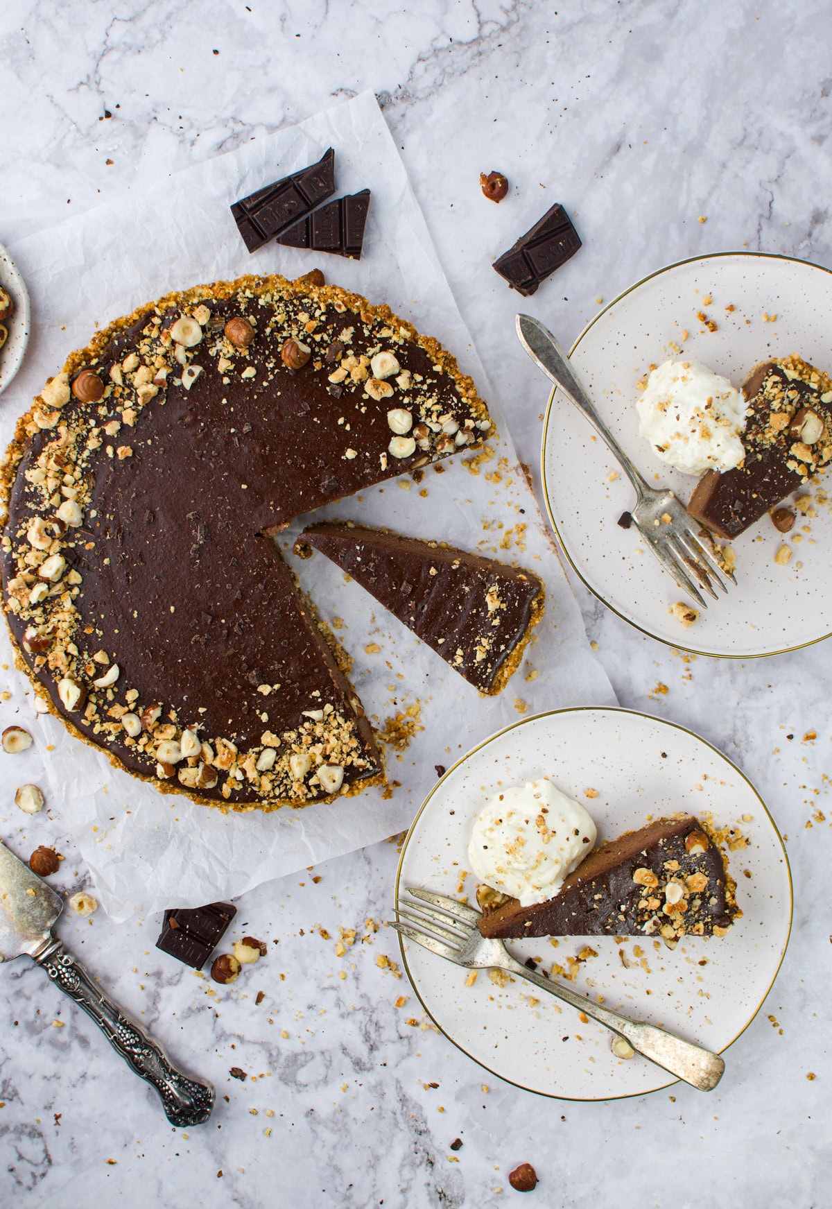 Overhead view of a Hazelnut Chocolate Tart with a slice removed, topped with chopped nuts. Two plates each hold a slice of tart and a scoop of ice cream. Pieces of chocolate and crumbs are scattered on a marble surface.