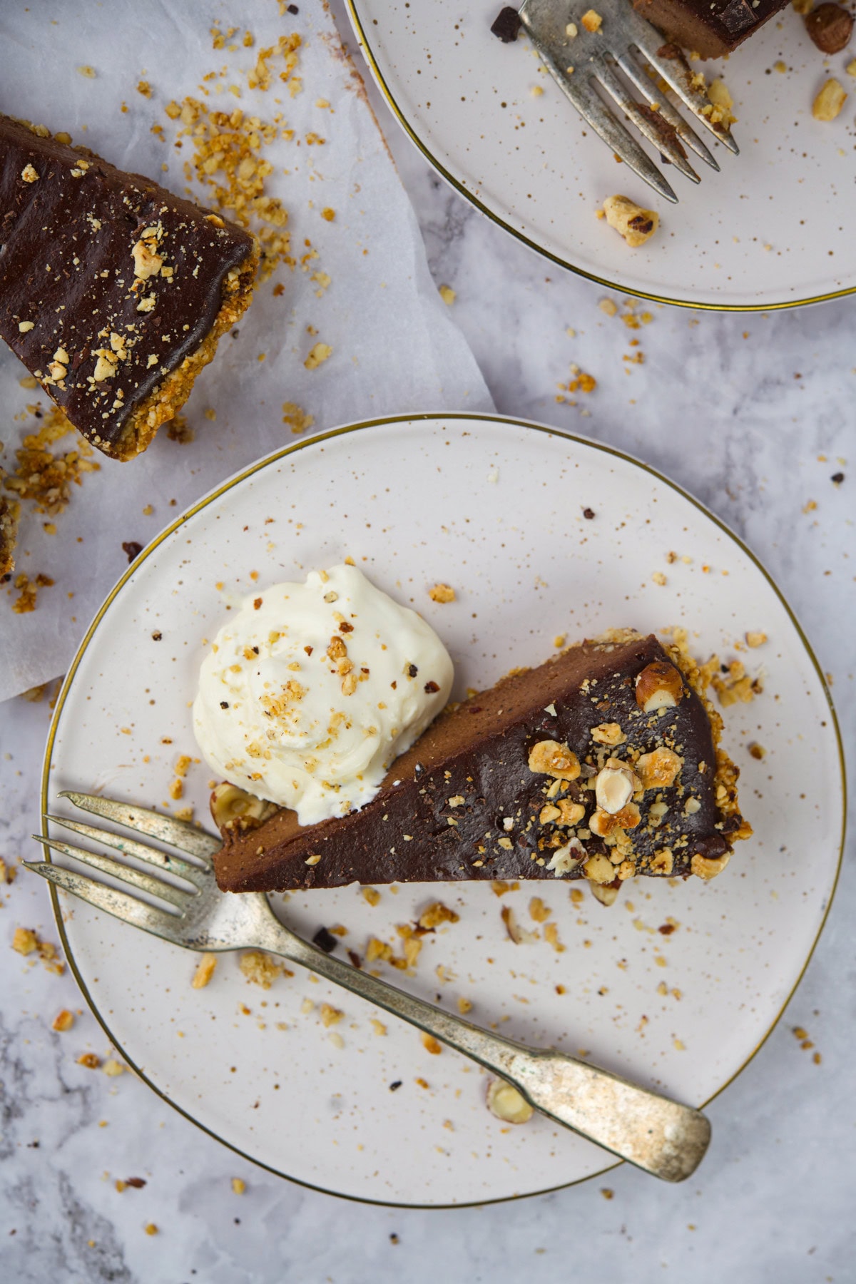 A slice of chocolate cake topped with crushed nuts, served with a dollop of whipped cream on a white plate with a gold rim. A fork rests on the plate, and crumbs are scattered around.
