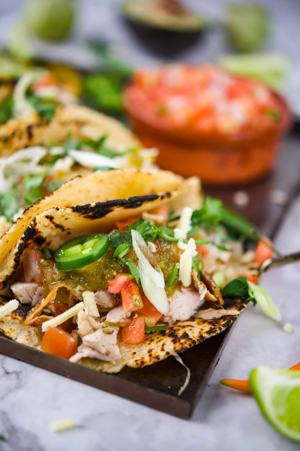Close-up of grilled chicken tacos in corn tortillas, topped with sliced jalape&ntilde;os, shredded cheese, diced tomatoes, and fresh herbs, with a bowl of salsa and lime wedges in the background.
