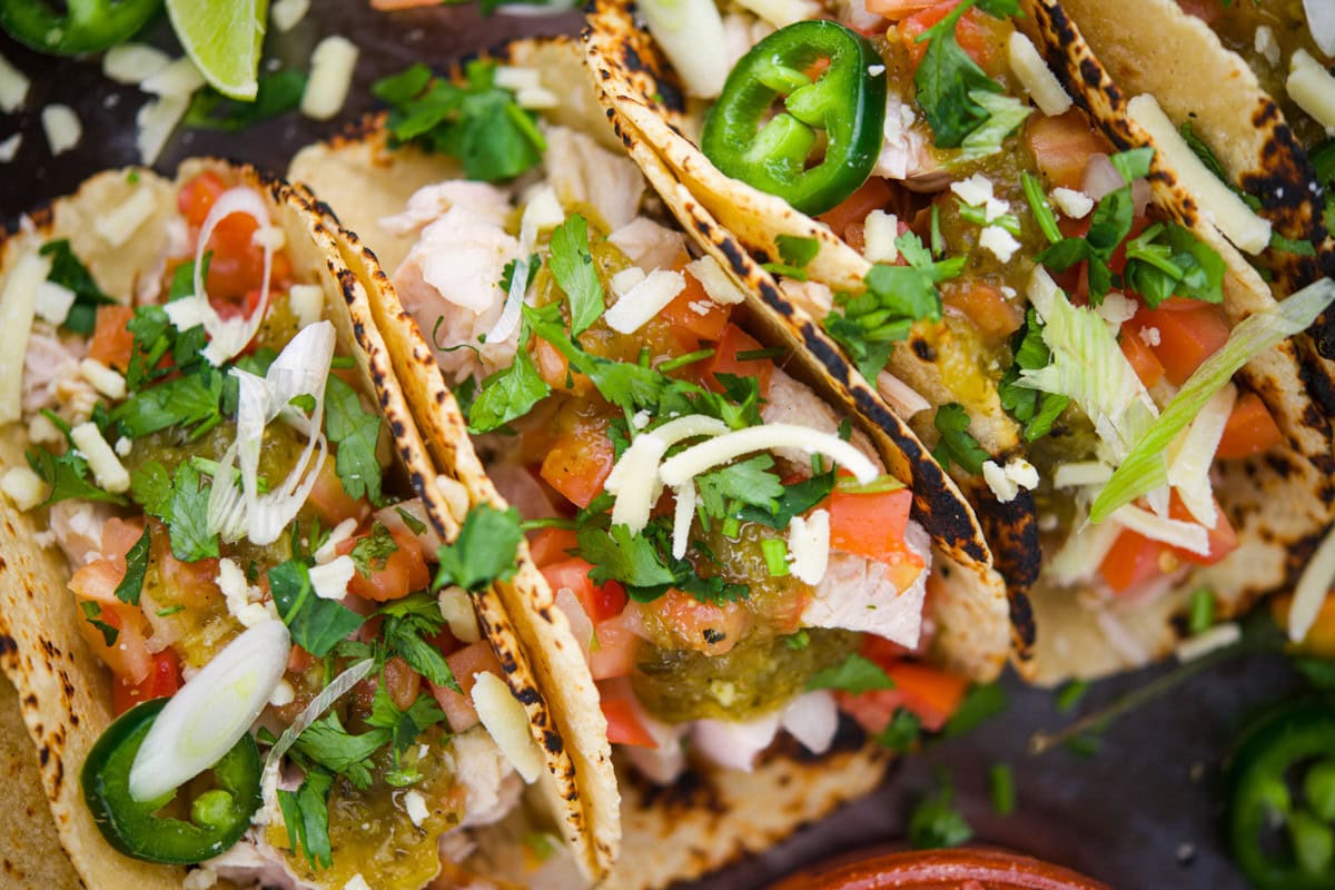 Three grilled tacos filled with diced chicken, chopped tomatoes, fresh cilantro, shredded cheese, sliced jalape&ntilde;os, green onions, and salsa, served on charred tortillas with lime wedges on the side.