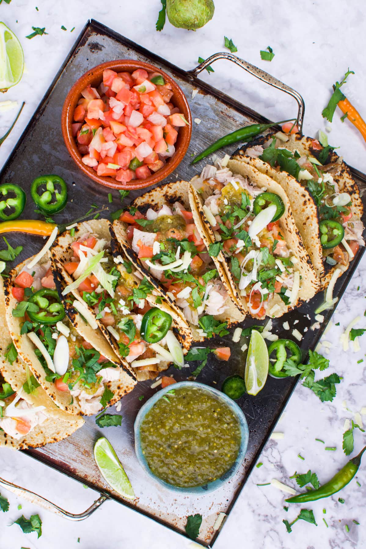 A tray of tacos filled with shredded meat, salsa, jalape&ntilde;os, cilantro, and cheese, garnished with lime wedges and served with bowls of salsa and pico de gallo on the side.