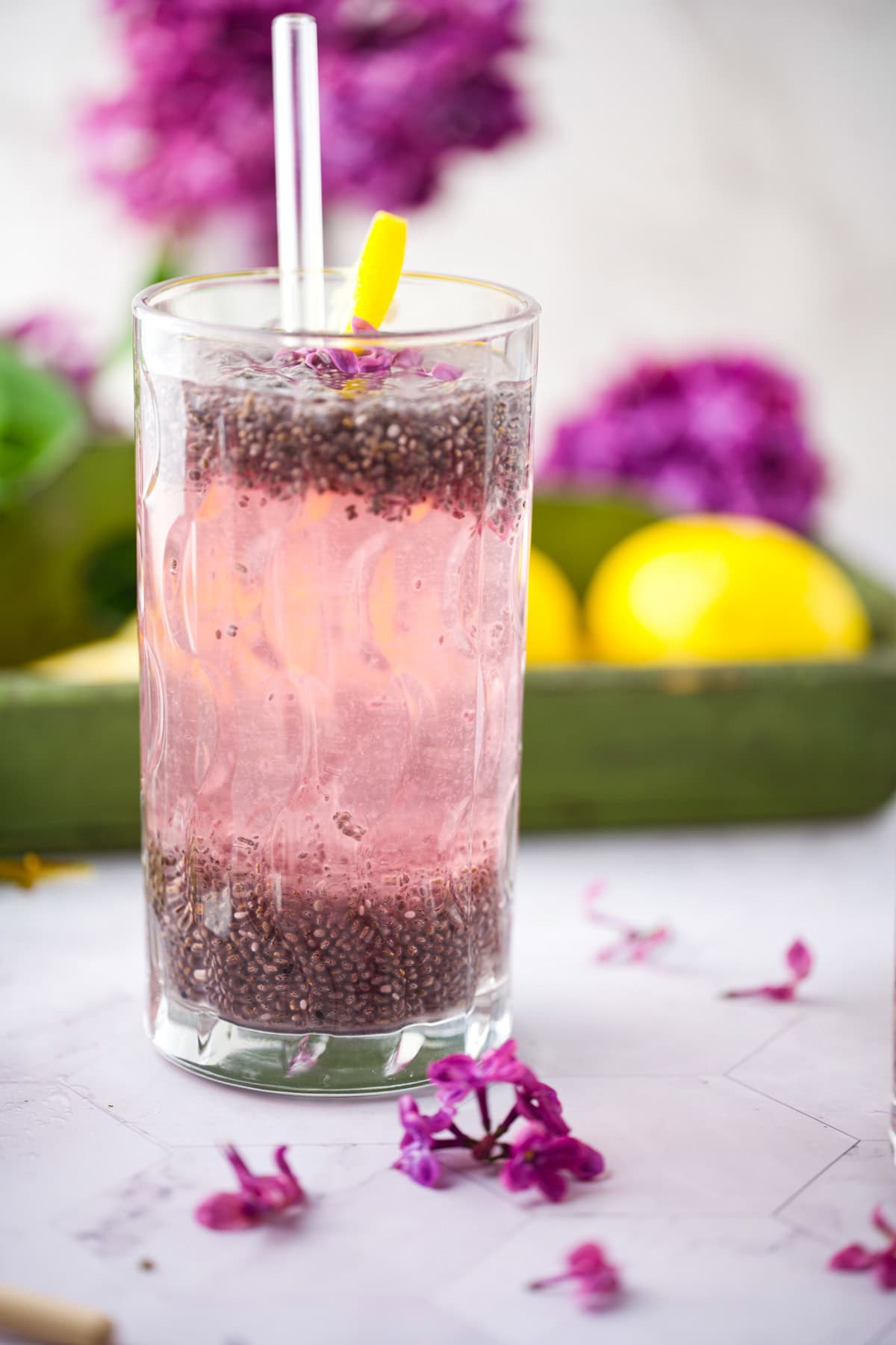 A tall glass of Lilac Chia Seed Lemonade drink with a clear straw and a lemon twist garnish, placed on a white surface with purple flowers and lemons in the background.