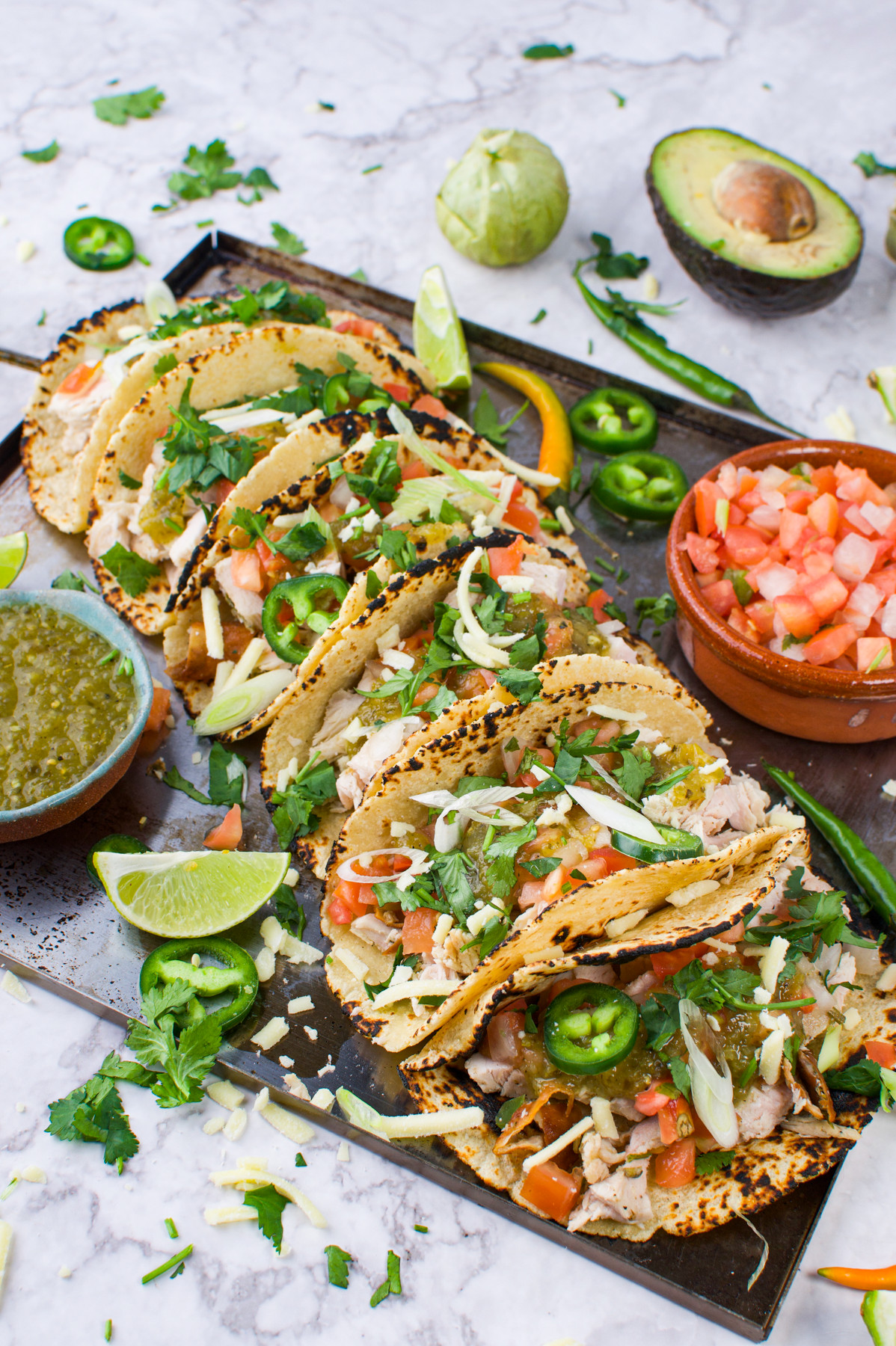 A tray of five Leftover Tureky Tacos, filled with shredded meat, lettuce, tomatoes, cheese, cilantro, and jalapeño slices, garnished with lime wedges. Salsa, avocado, and green sauce are nearby on a marble surface.
