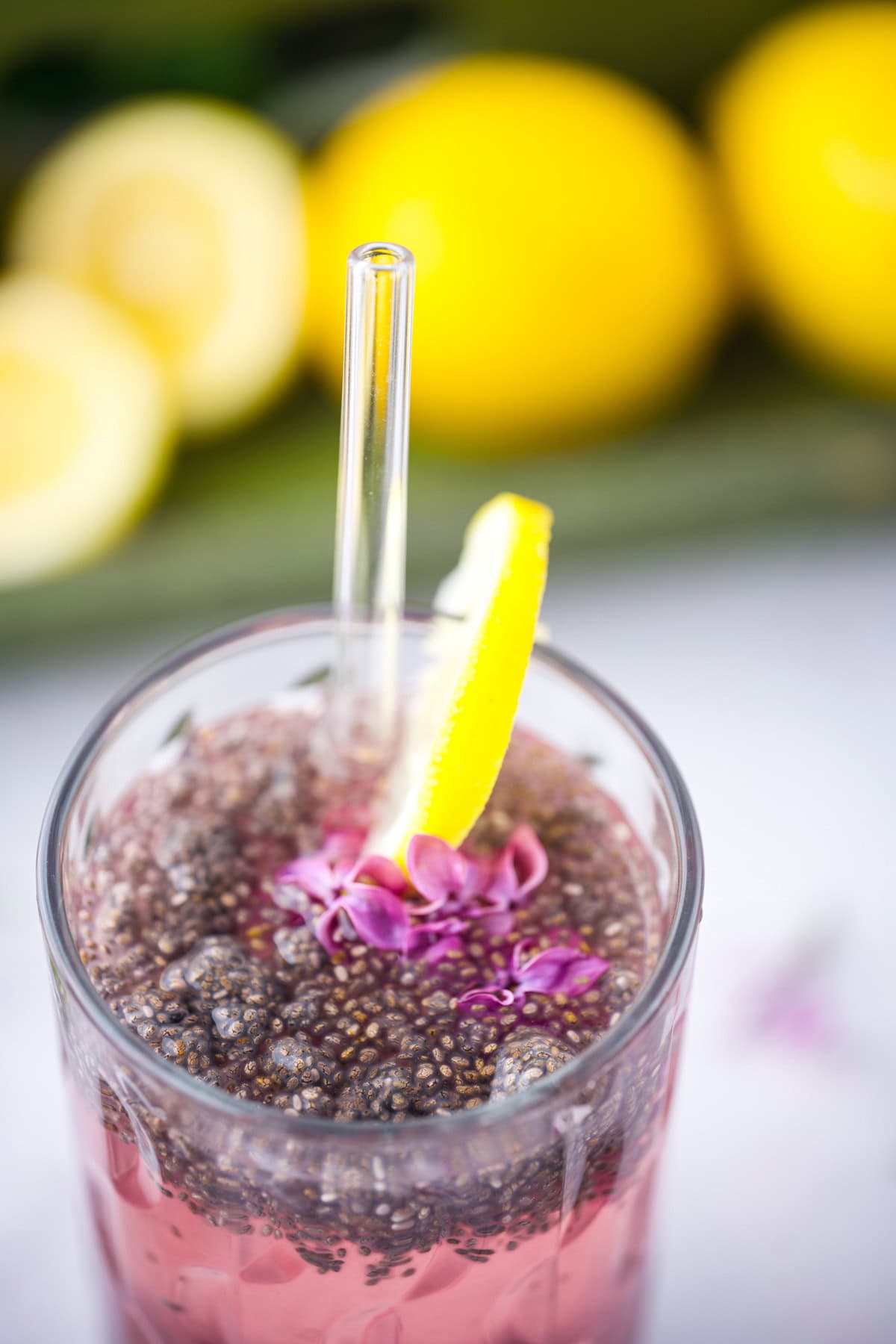 A close-up of a pink chia seed drink in a glass with a clear straw, garnished with a lemon slice and purple edible flowers, with whole and sliced lemons blurred in the background.