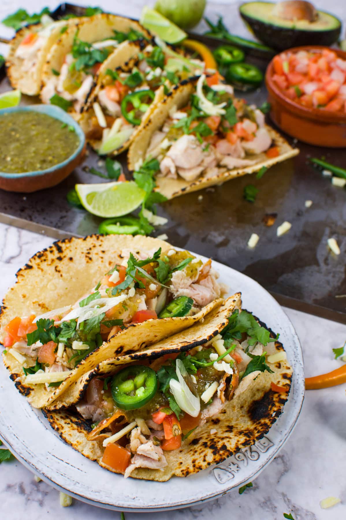 A plate of grilled chicken tacos topped with diced tomatoes, onions, jalape&ntilde;os, shredded cheese, and cilantro, with lime wedges and bowls of salsa on the side. More tacos are arranged on a tray in the background.