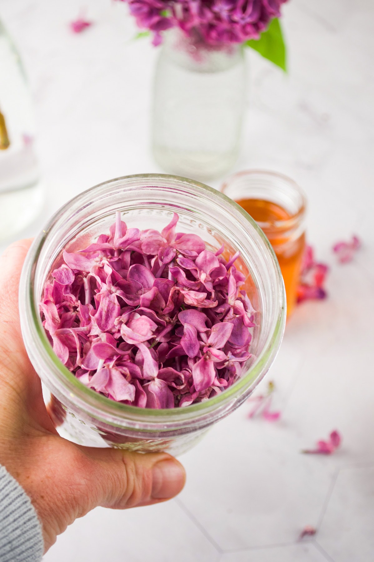 A hand holds a glass jar filled with purple flower petals. In the background, jars with orange liquid and more flowers are visible on a white surface.