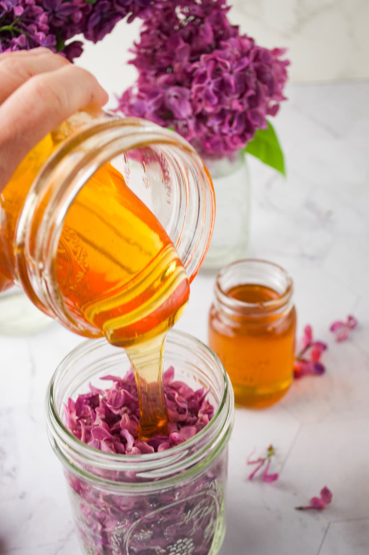 A hand pours golden honey from a jar into another jar filled with fresh purple lilac flowers, with more lilacs and a small jar of honey in the background on a white marble surface.
