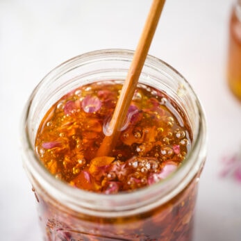 A close-up of a glass jar filled with Lilac Flower Infused Honey being stirred with a wooden spoon.