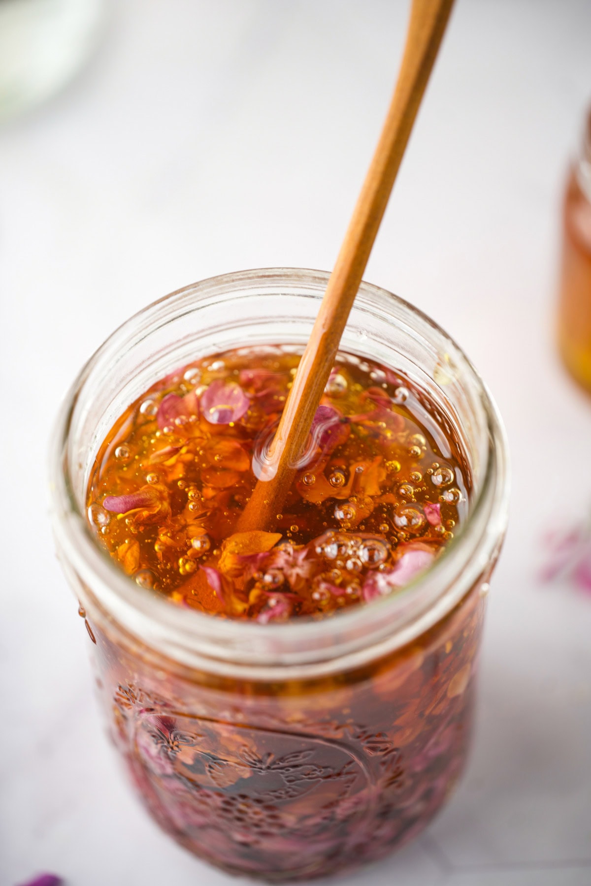 A close-up of a glass jar filled with Lilac Flower Infused Honey being stirred with a wooden spoon.