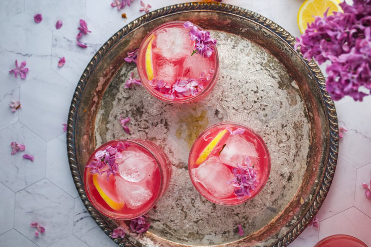 Three glasses with pink drinks, ice cubes, lemon slices, and purple flowers sit on an ornate silver tray, with flower petals and lemons scattered on a white marble surface.