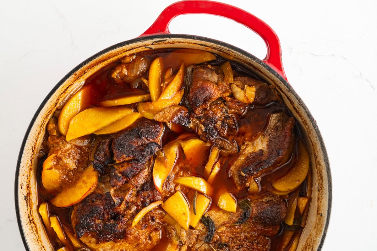 A red Dutch oven filled with apple cider braised pork shoulder and sliced apples in a rich, brown sauce, all set against a white background.