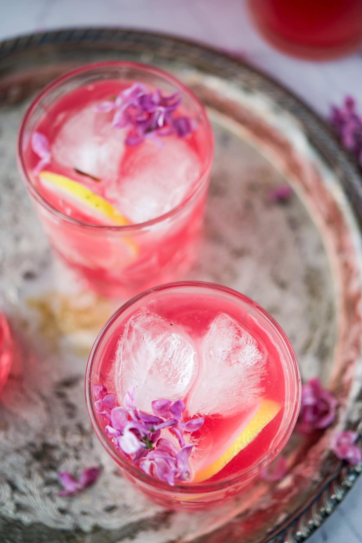 Two glasses of pink iced drinks garnished with purple flowers and lemon slices sit on a rustic silver tray. The drinks are bright and refreshing, with large ice cubes and delicate floral decorations.