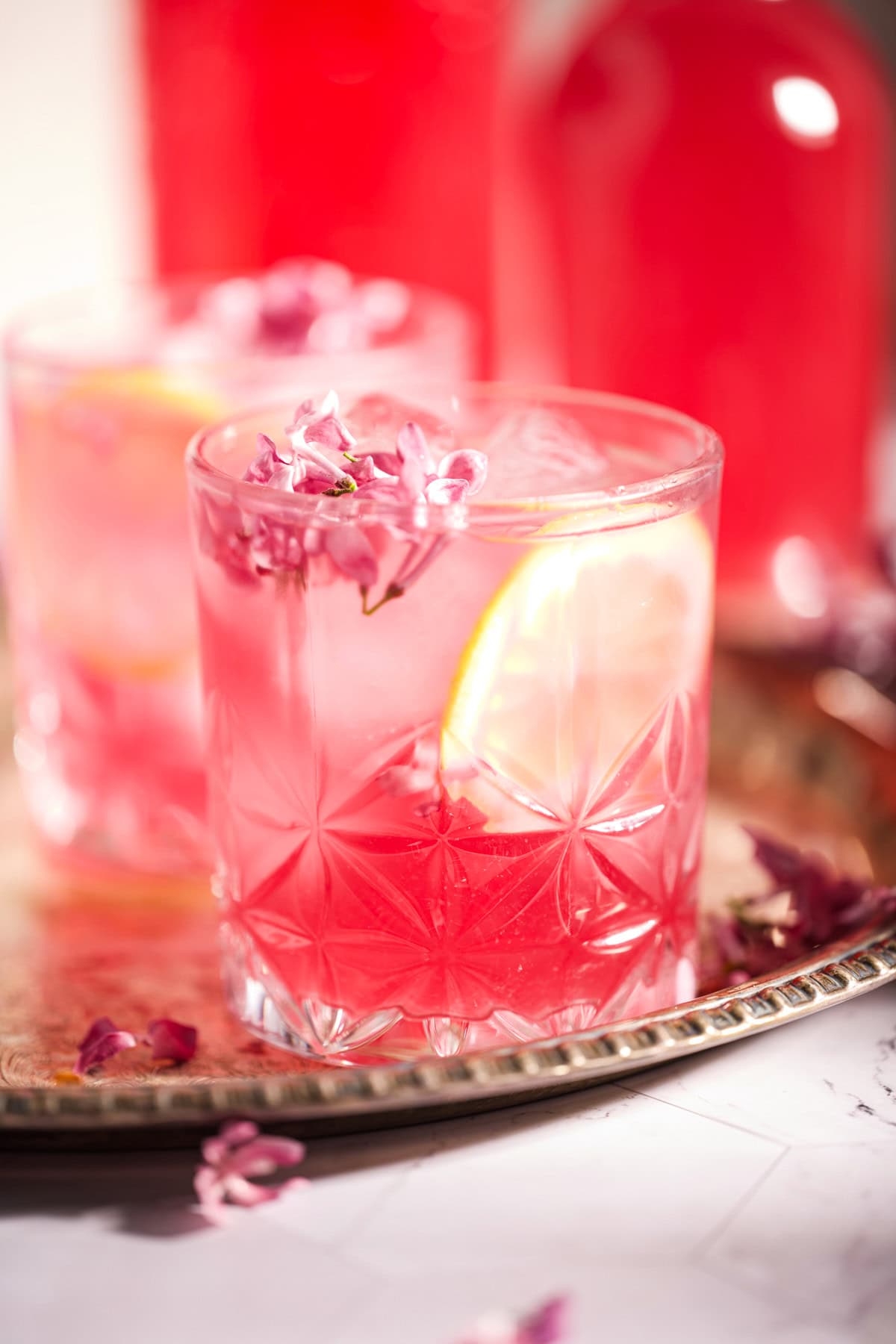 A glass of Fermented Lilac Soda with ice, a lemon slice, and edible flowers on top sits on a silver tray; another similar glass and a bottle are blurred in the background.