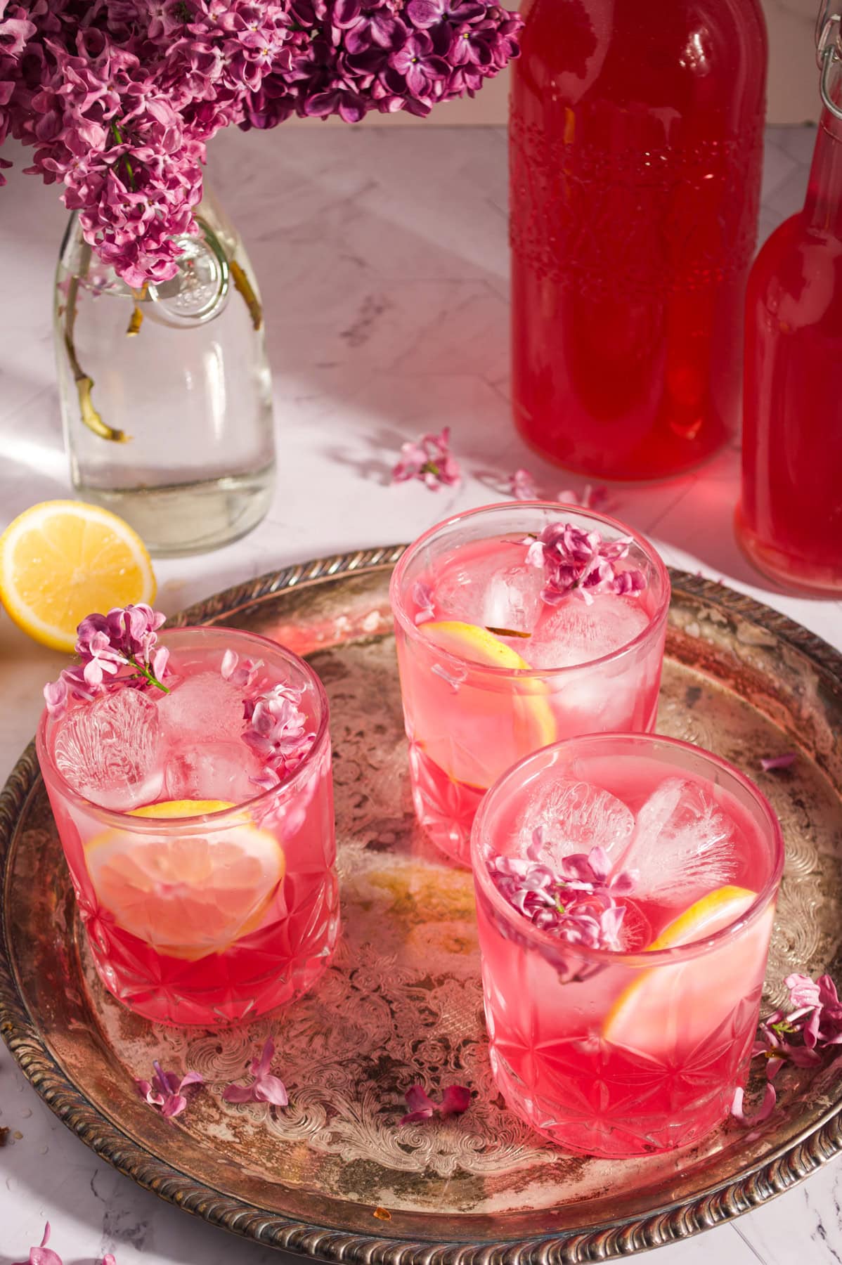 Three glasses of pink lemonade with ice, lemon slices, and purple flower garnishes sit on a silver tray. A bottle of pink drink, a lemon half, and a bouquet of lilacs are in the background.