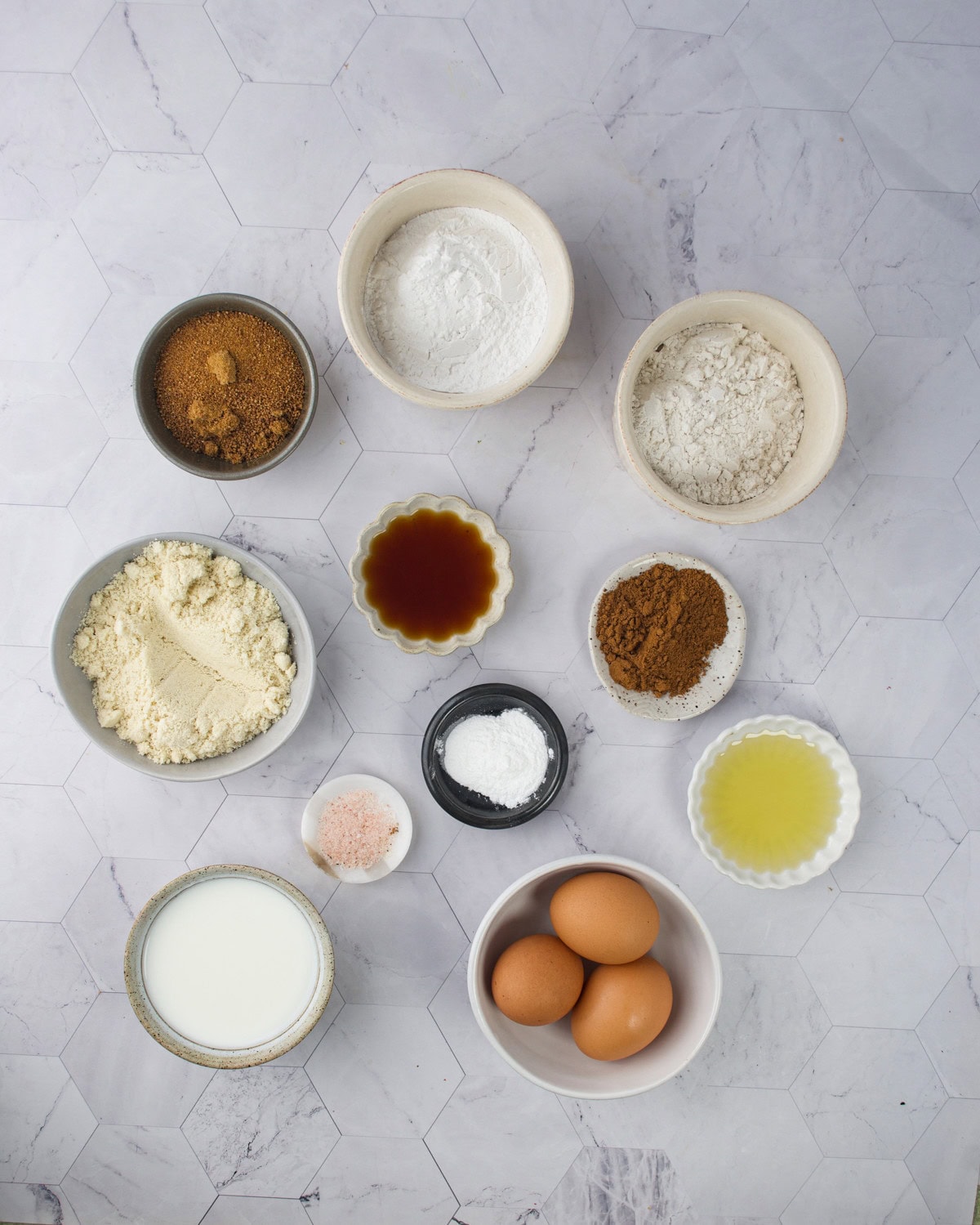 A top-down view of bowls and dishes containing baking ingredients: flour, almond flour, cocoa powder, sugar, salt, eggs, milk, baking powder, vanilla extract, and oil on a light hexagon-patterned surface.