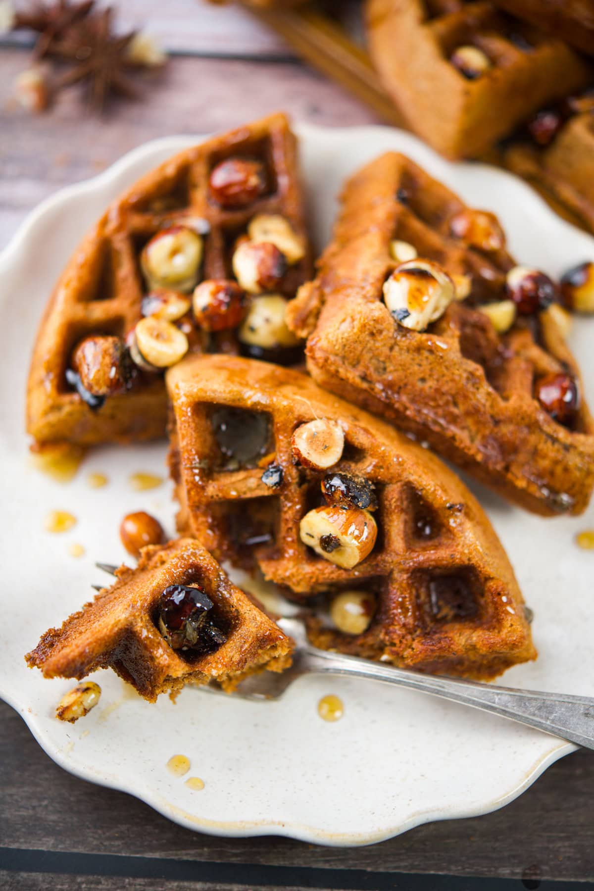 A plate with four pieces of waffle, topped with whole toasted hazelnuts and drizzled with syrup. A fork holds a bite-size piece of waffle. The background shows a rustic wooden table.