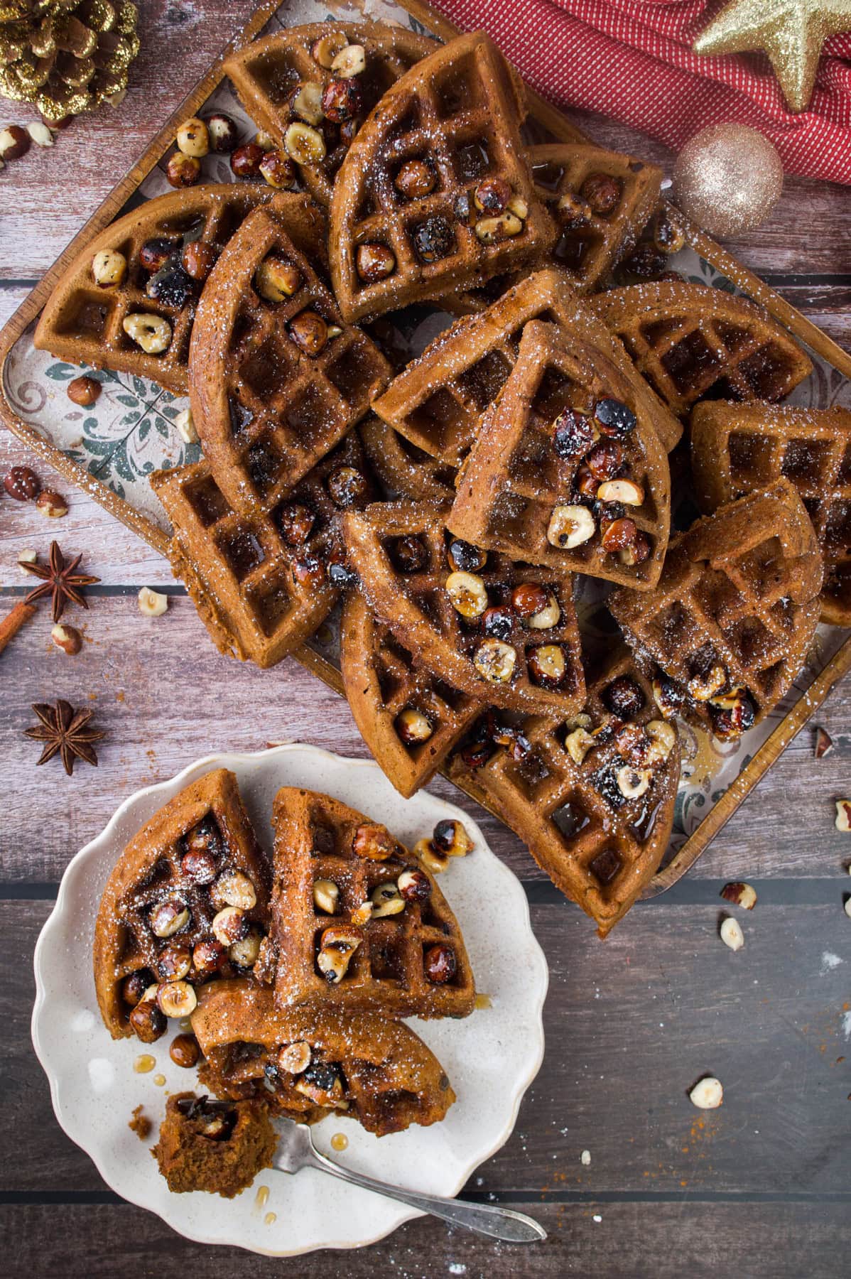A plate with sliced waffles topped with hazelnuts and dried fruit sits beside a tray with more waffles on a rustic wooden table, surrounded by festive decorations and spices.