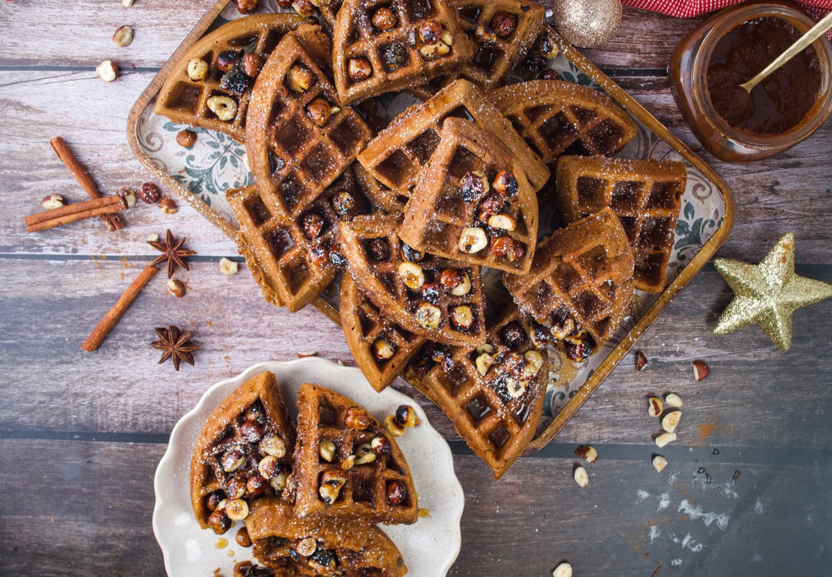 A tray and plate filled with sliced waffles topped with chopped hazelnuts and powdered sugar, next to a jar of chocolate sauce, cinnamon sticks, and festive holiday decorations on a wooden table.