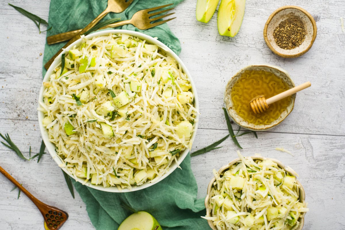 A bowl of fresh apple and celery root slaw sits on a green cloth, surrounded by lemon slices, honey, pepper, and utensils on a rustic white table. A smaller bowl of slaw is nearby, garnished with fresh herbs.