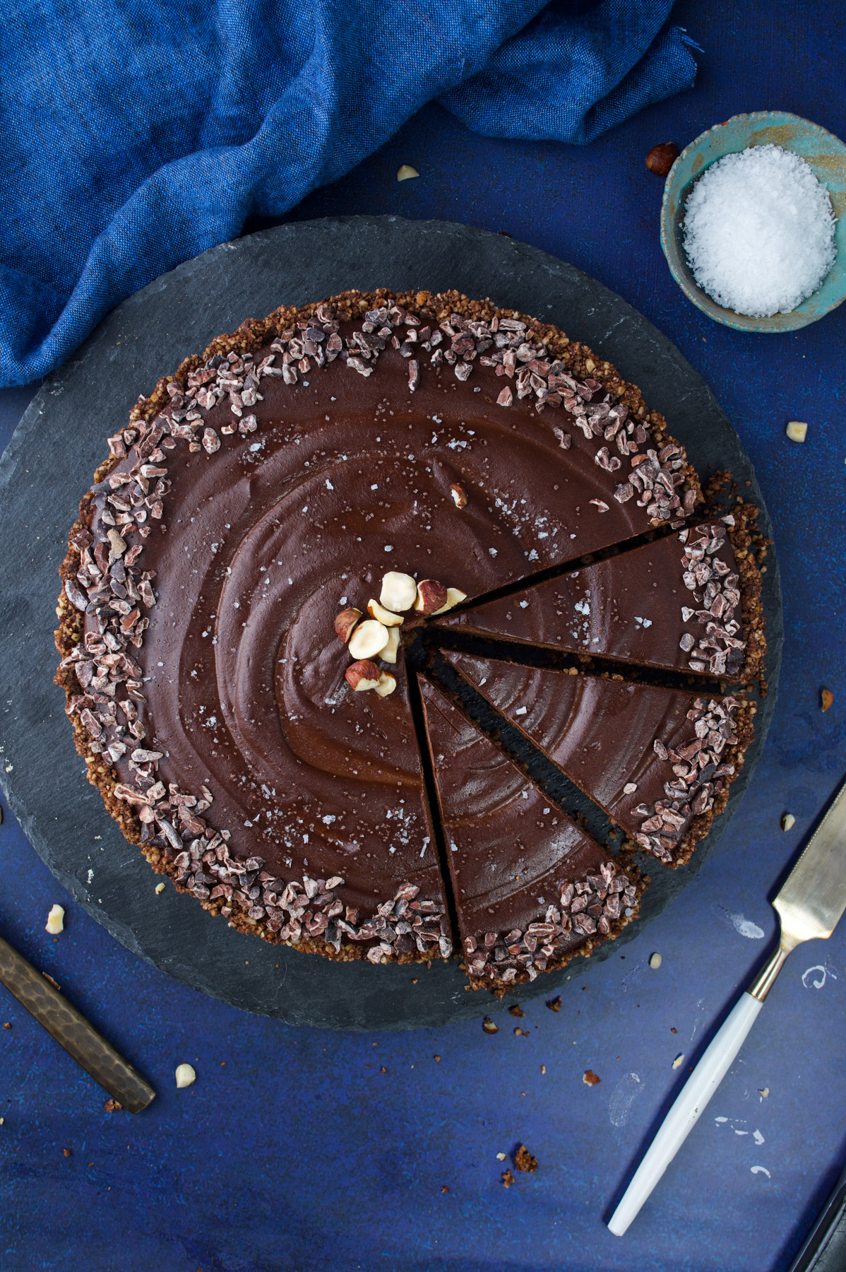 A no-bake chocolate tart with a smooth, glossy surface, topped with chopped hazelnuts, cocoa nibs, and a sprinkle of sea salt. Two slices are cut and slightly separated. A pie server and bowl of salt are nearby on a dark blue background.