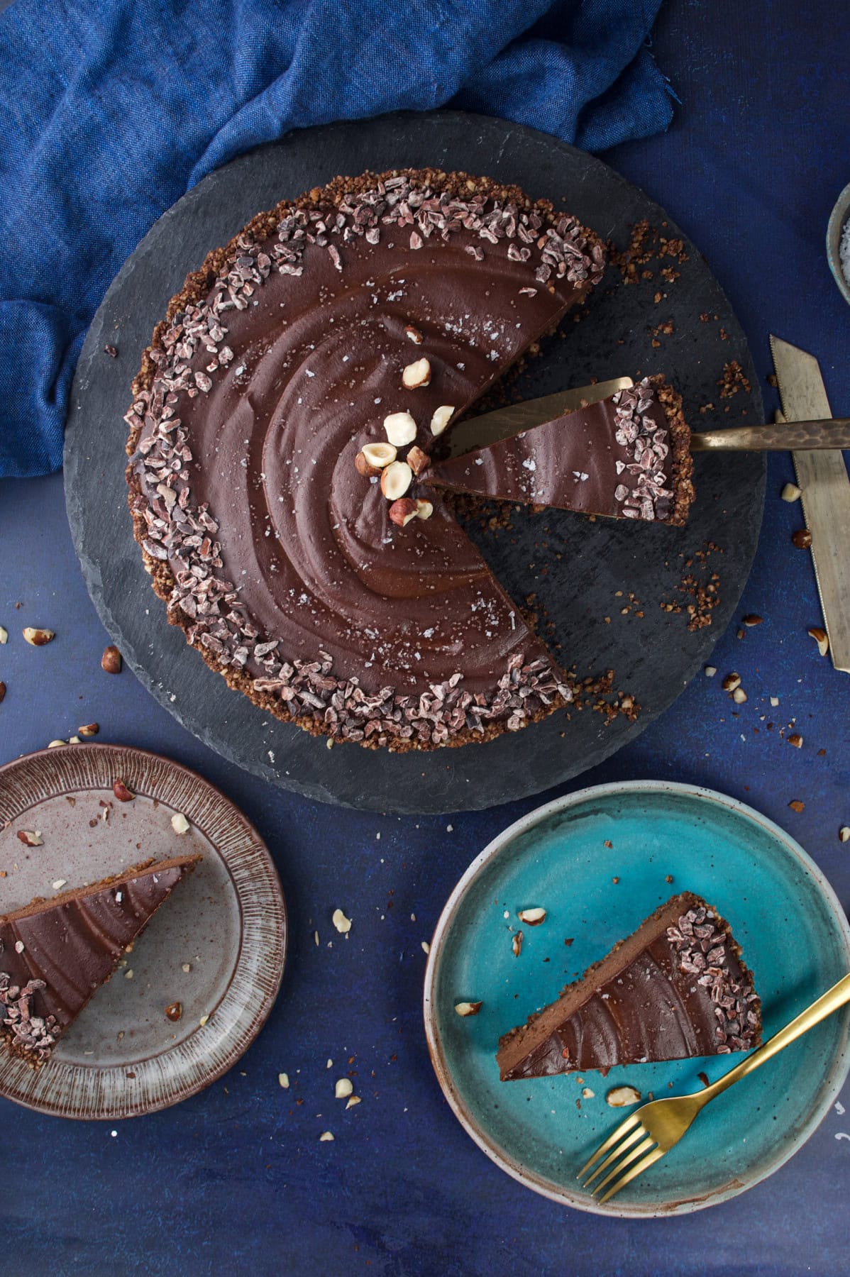 A sliced chocolate tart topped with chopped nuts sits on a dark slate board. One slice is being served; two plates with slices are nearby, along with a gold fork. Blue fabric is in the background.