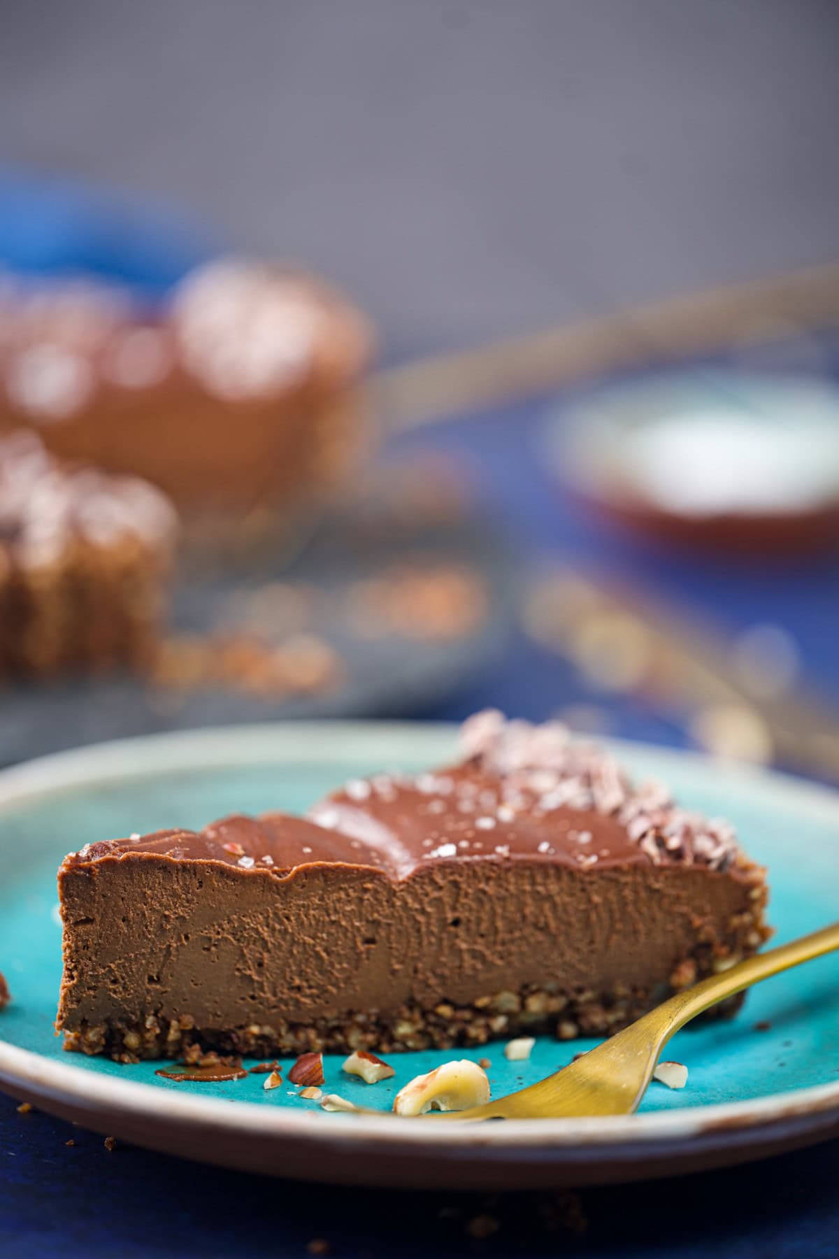 A close-up of a slice of chocolate cheesecake on a turquoise plate with a gold fork, showing a creamy texture and nutty crust, with more cheesecake blurred in the background.