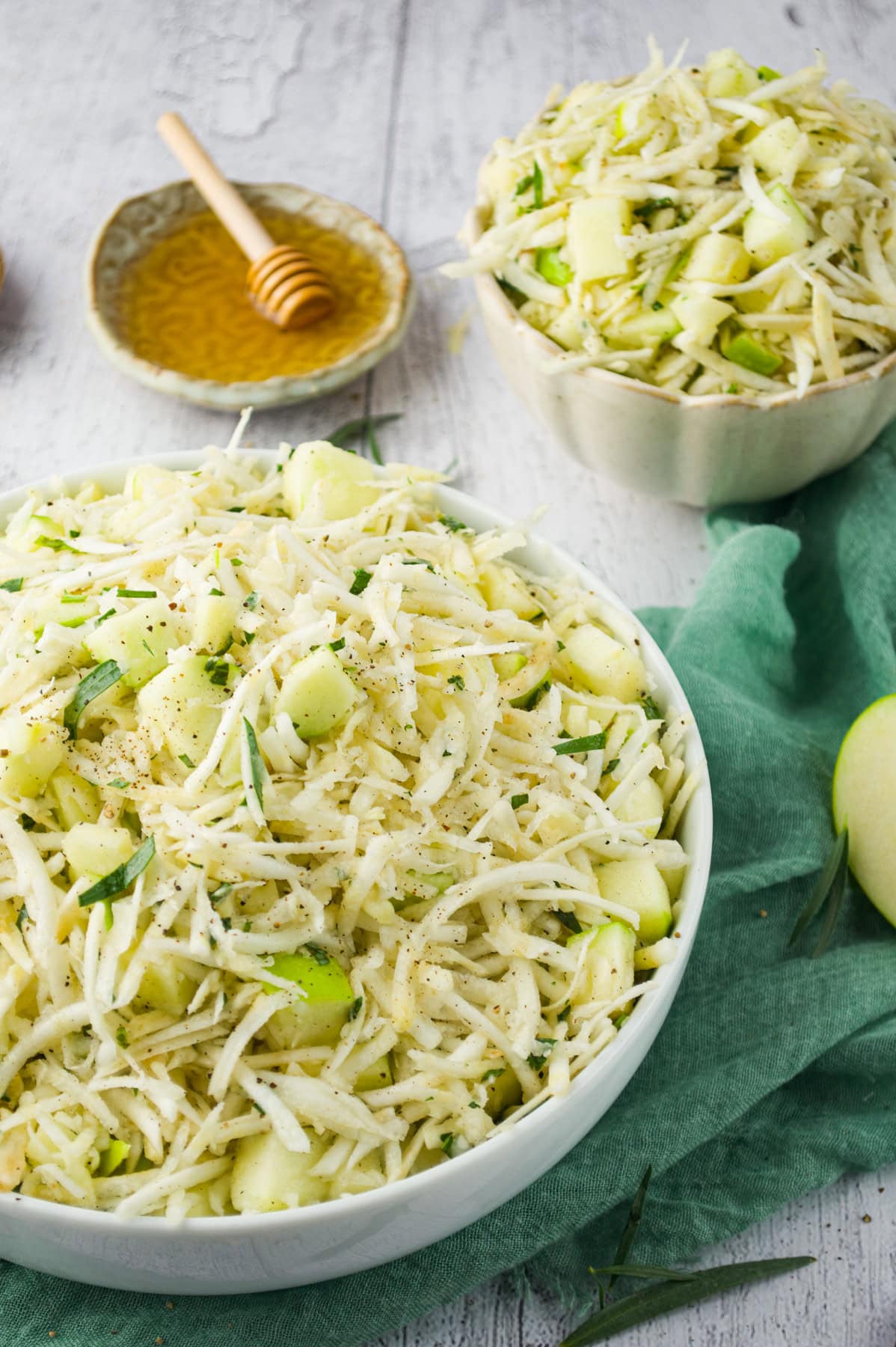 Two bowls of shredded apple and celery root salad garnished with herbs are on a wooden table, with a green cloth and a small dish of honey with a honey dipper in the background.