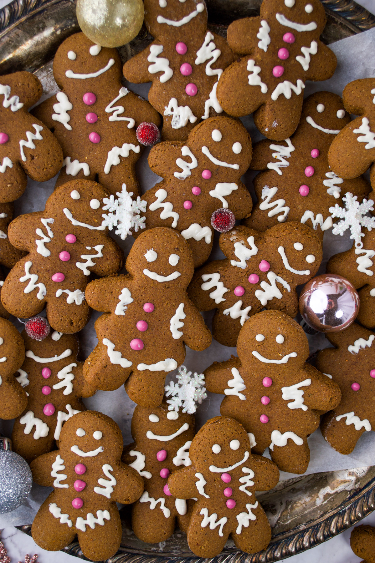 A tray filled with decorated gingerbread cookies shaped like people, featuring white icing details and pink buttons, surrounded by decorative ornaments and snowflake sprinkles.