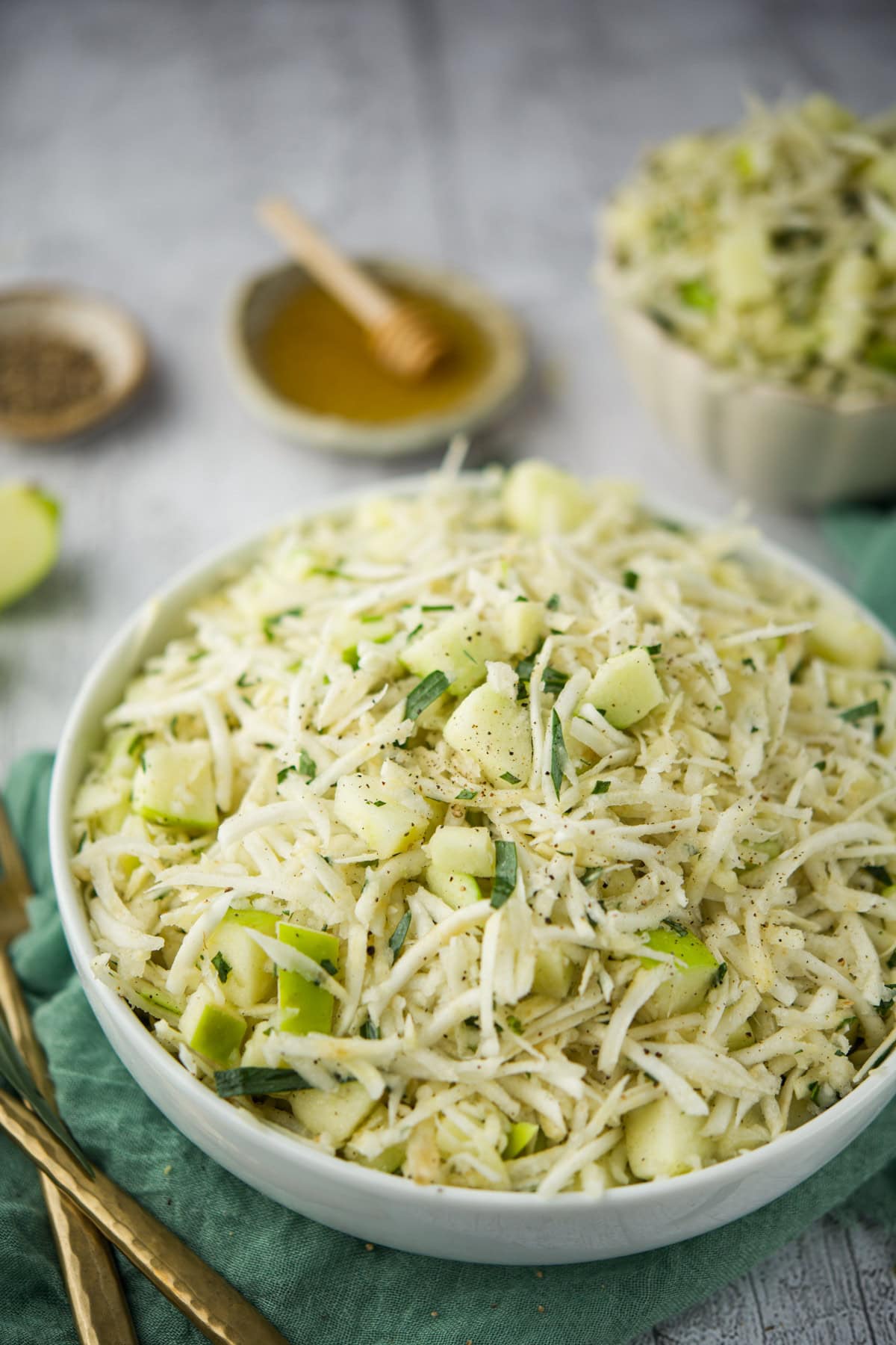 A bowl of fresh salad featuring shredded celeriac, diced green apple, and herbs, sprinkled with black pepper. In the background, a small dish of honey and a bowl of salad are visible on a rustic surface.