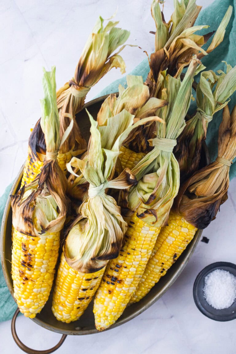Grilled corn on the cob with charred husks, neatly arranged in a rustic copper pan on a white surface with a green cloth