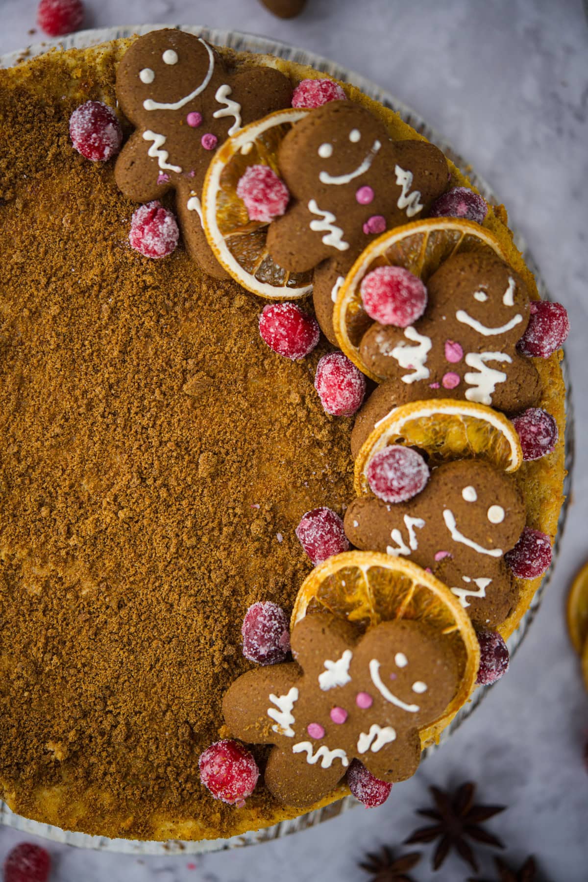 A pie topped with gingerbread cookies shaped like people, sugared cranberries, and dried orange slices, arranged on one side; the rest of the pie has a smooth, crumbly brown surface.