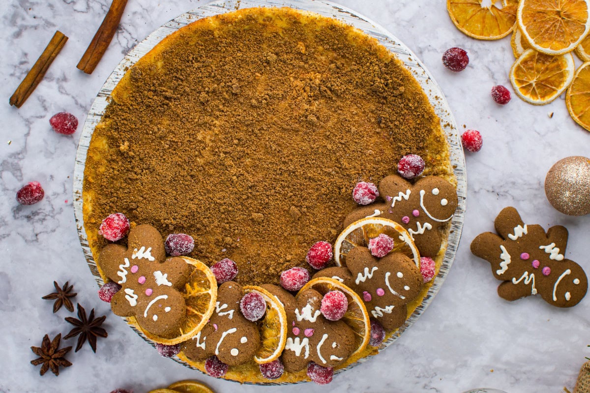 A Gingerbread Cheesecake topped with a crumbly brown layer, decorated with sugared cranberries, dried orange slices, and gingerbread cookies shaped like people on a marble surface with festive spices and decorations scattered around.