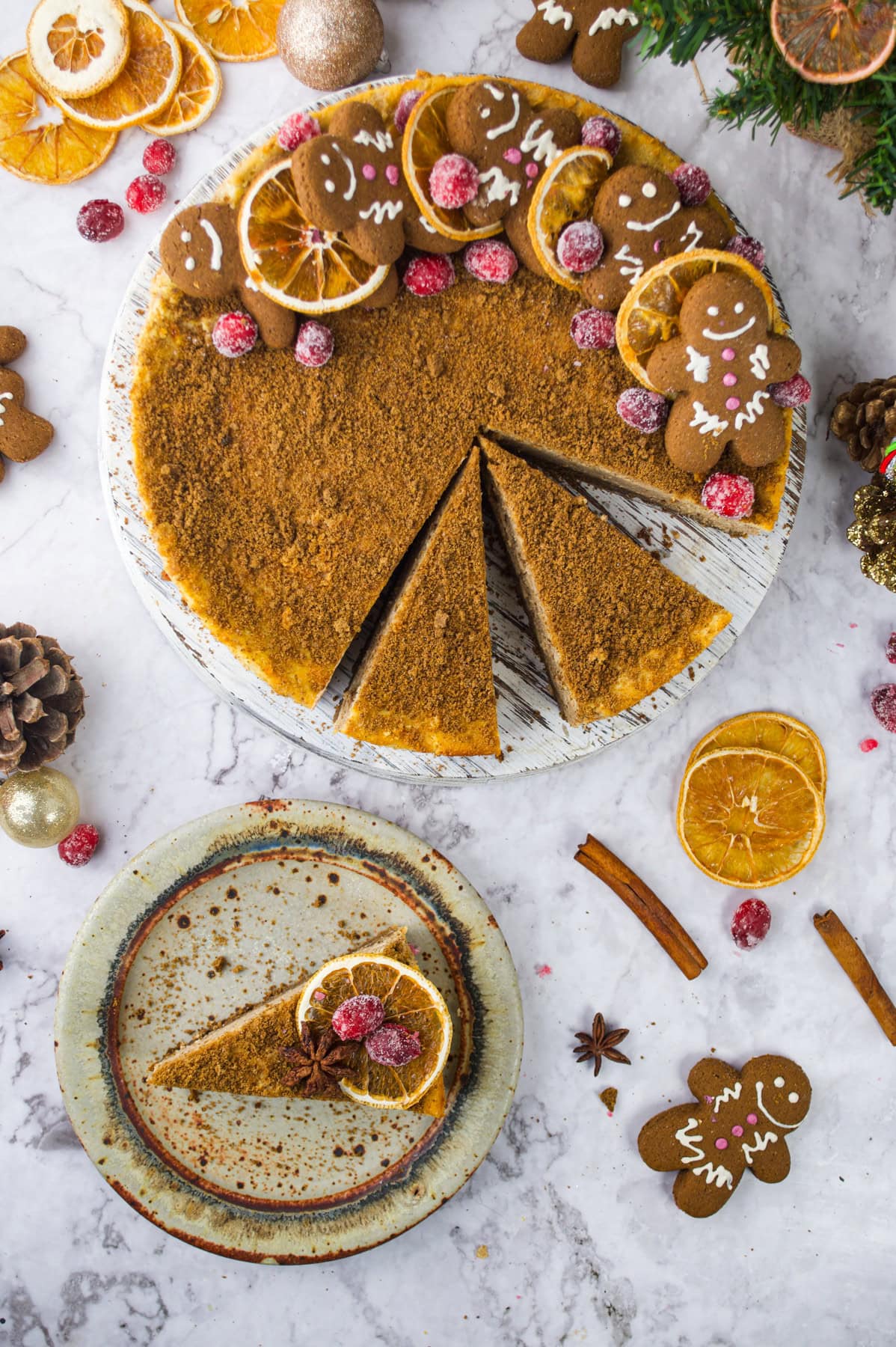 A festive cheesecake topped with gingerbread cookies and sugared cranberries, with one slice on a plate garnished with dried orange slices and cranberries. The scene includes holiday decorations on a marble surface.