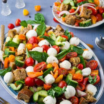 Colorful panzanella salad on a decorative plate, featuring chunks of crusty bread, cherry tomatoes, sliced cucumbers, yellow bell peppers, fresh basil leaves, red onion, and mozzarella balls, with a gold fork beside the plate and a blurred background