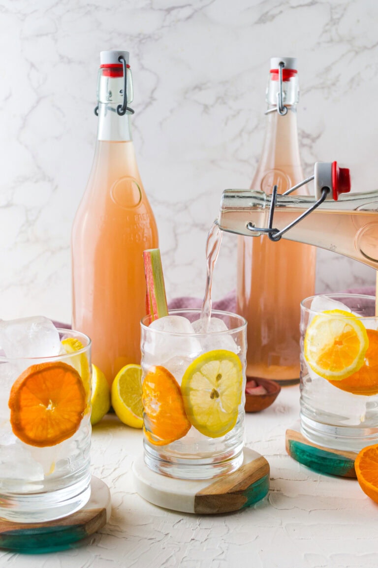 A hand pours a light pink fermented soda from a glass flip-top bottle into a tumbler filled with ice, garnished with slices of orange, lemon, and a stalk of rhubarb. More bottles of the soda are visible in the background on a light marble surface.