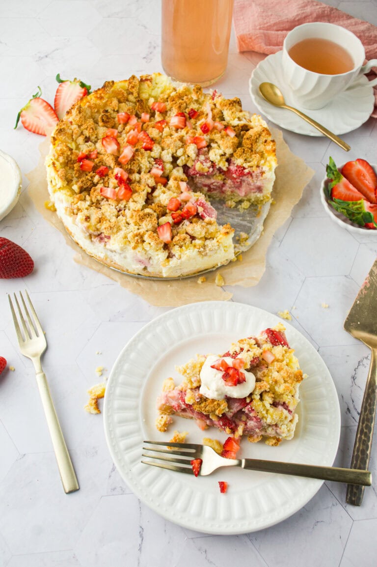 A sliced strawberry and rhubarb crumb cake on parchment paper, topped with fresh strawberries, served with tea and a plated slice garnished with whipped cream and berries