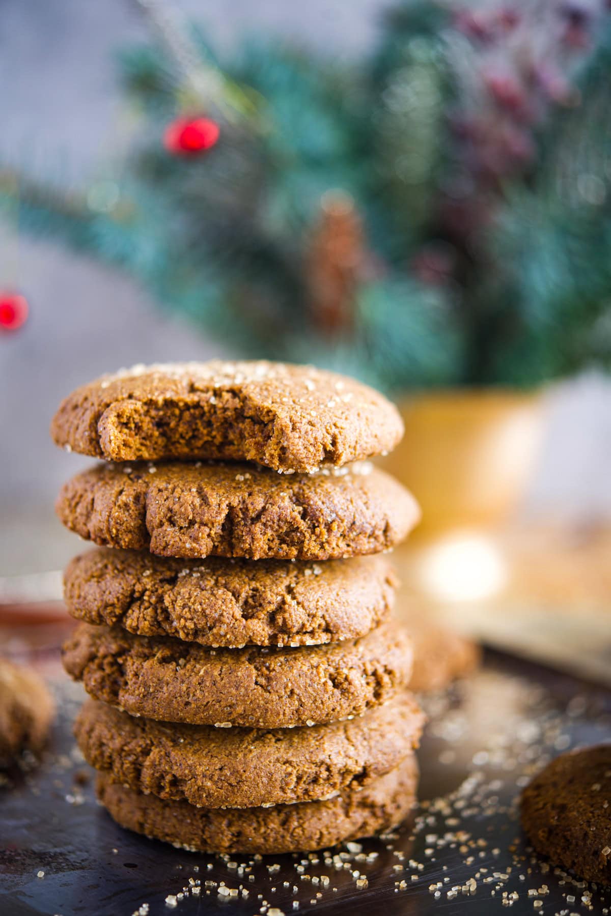 A stack of six Gluten-Free Molasses Cookies, with the top cookie having a bite taken out of it, sits on a dark surface. In the blurred background, there is a festive arrangement of pine branches and red berries.