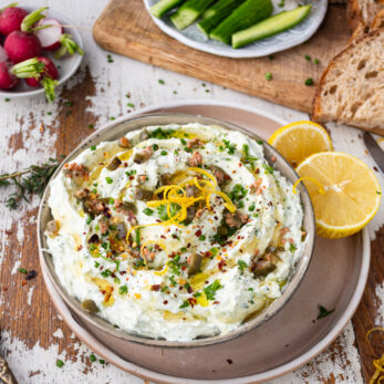 A bowl of Mediterranean Spring Herb Whipped Feta Dip garnished with lemon zest, chives, and chili flakes, surrounded by lemon halves. Nearby are bread slices, radishes, cucumber slices, and more lemon wedges on a wooden board.