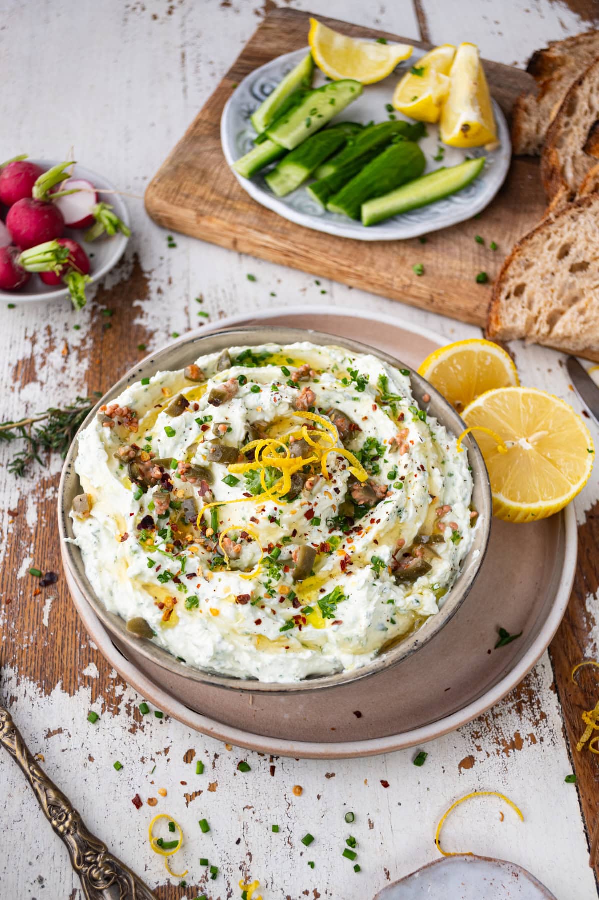 A bowl of creamy dip garnished with lemon zest, chives, and chili flakes, surrounded by lemon halves. Nearby are bread slices, radishes, cucumber slices, and more lemon wedges on a wooden board.