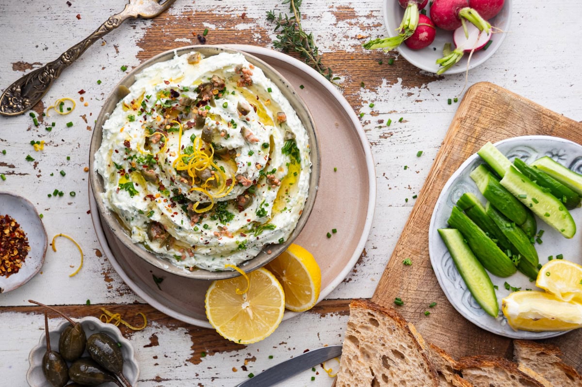 A bowl of creamy dip topped with herbs, lemon zest, and olive oil sits on a plate with lemon wedges. Surrounding it are cucumbers, radishes, bread, capers, and red pepper flakes on a rustic wooden table.