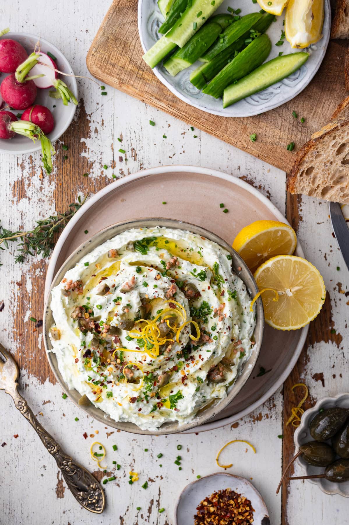A bowl of creamy whipped feta dip garnished with herbs and lemon zest sits on a plate, surrounded by lemon slices, fresh bread, cucumber sticks, radishes, and small dishes of spices and capers on a rustic wooden table.