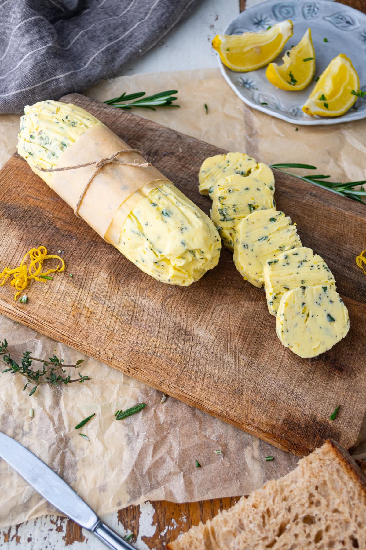 A log of herb butter, partially wrapped in parchment, sits on a wooden cutting board. Several round slices are cut. Fresh herbs, lemon wedges, and bread are nearby.