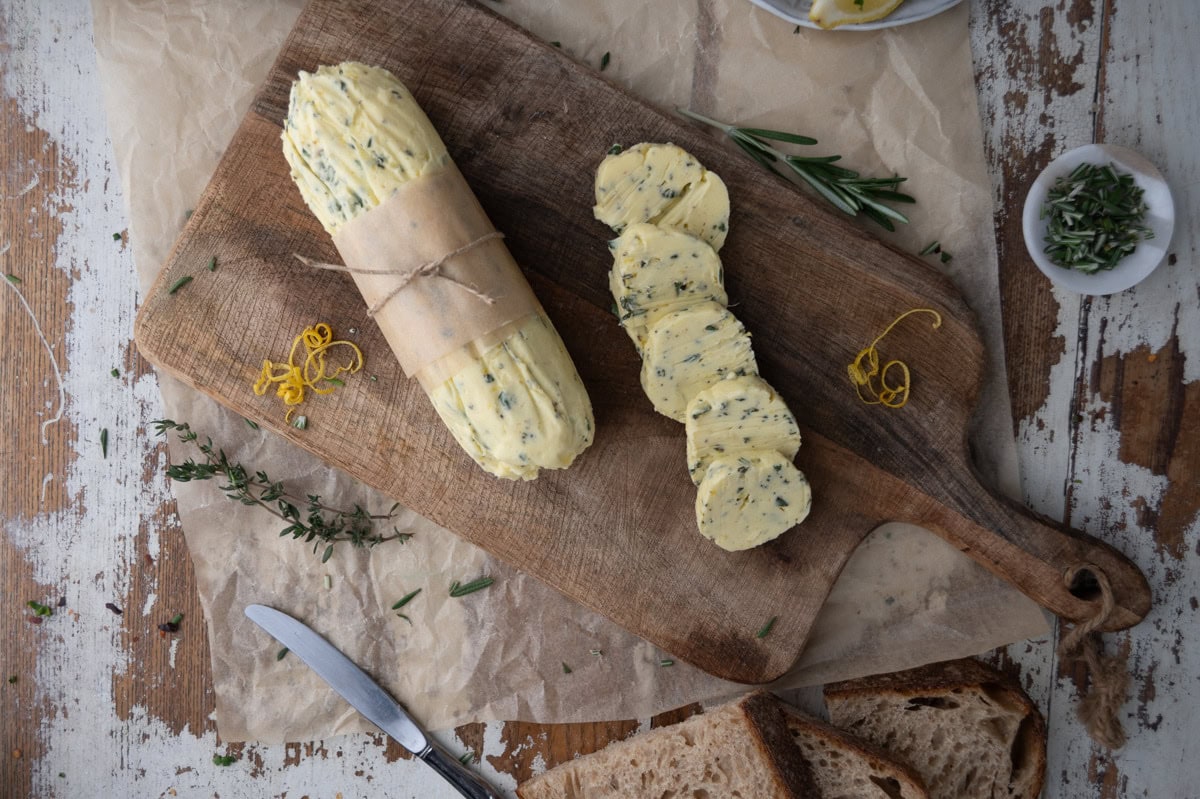 A log of herb butter wrapped in parchment, partially sliced, sits on a wooden cutting board with scattered herbs and lemon zest. A knife, bread, and small bowls of herbs are nearby on a rustic table.