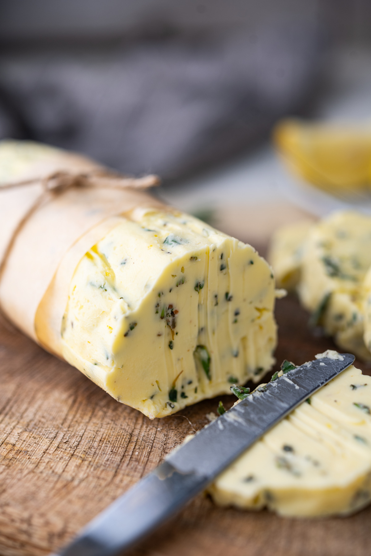 A close-up of a log of herb butter wrapped in parchment paper on a wooden board, with slices cut and a knife resting nearby. Fresh green herbs are visible in the creamy butter.