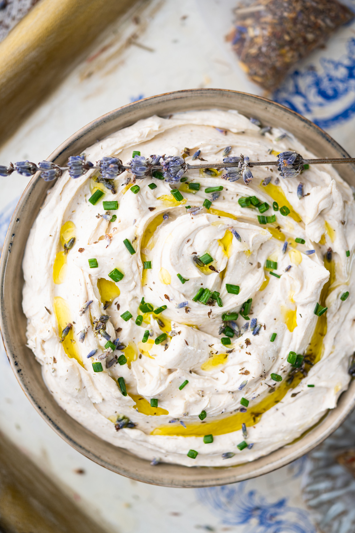 A bowl of Lavender Lemon-Infused Cream Cheese Dip garnished with chopped chives, dried herbs, a drizzle of olive oil, and a sprig of lavender, presented on a decorative plate.