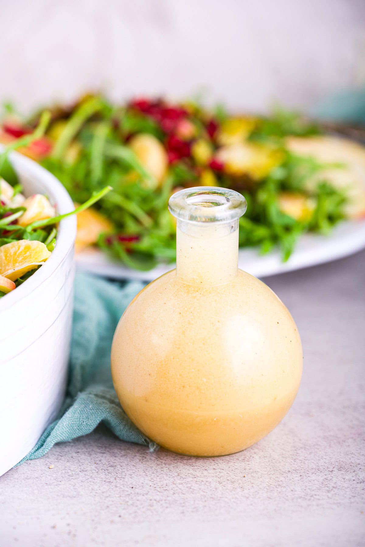 Vegan salad dressing in a glass bottle with fresh greens and fruit salad in the background.