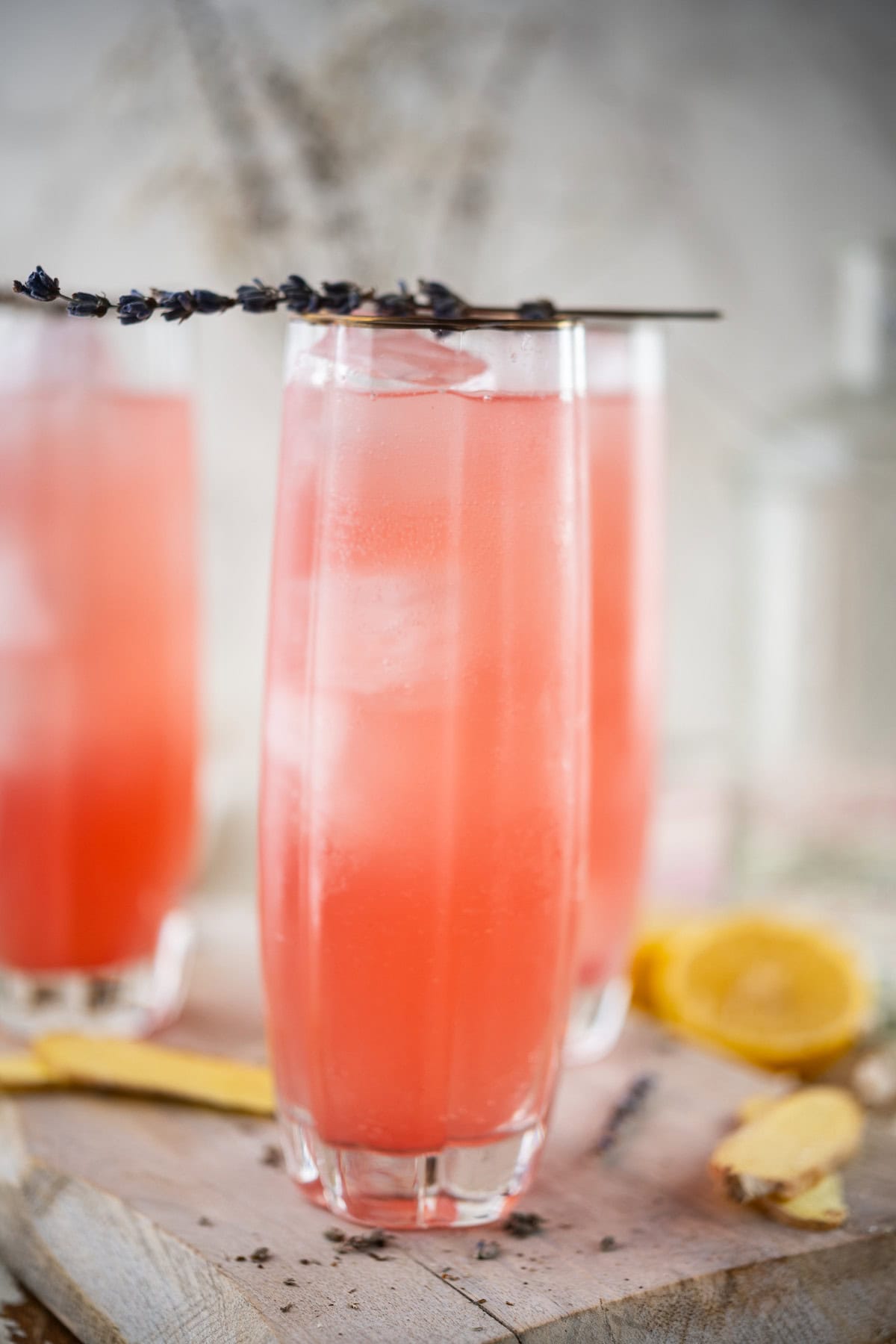 Tall glasses filled with Rhubarb Lavender And Hibiscus Cooler, are garnished with sprigs of lavender. Lemon slices and ginger pieces rest on a wooden surface in the foreground, with a blurred background.