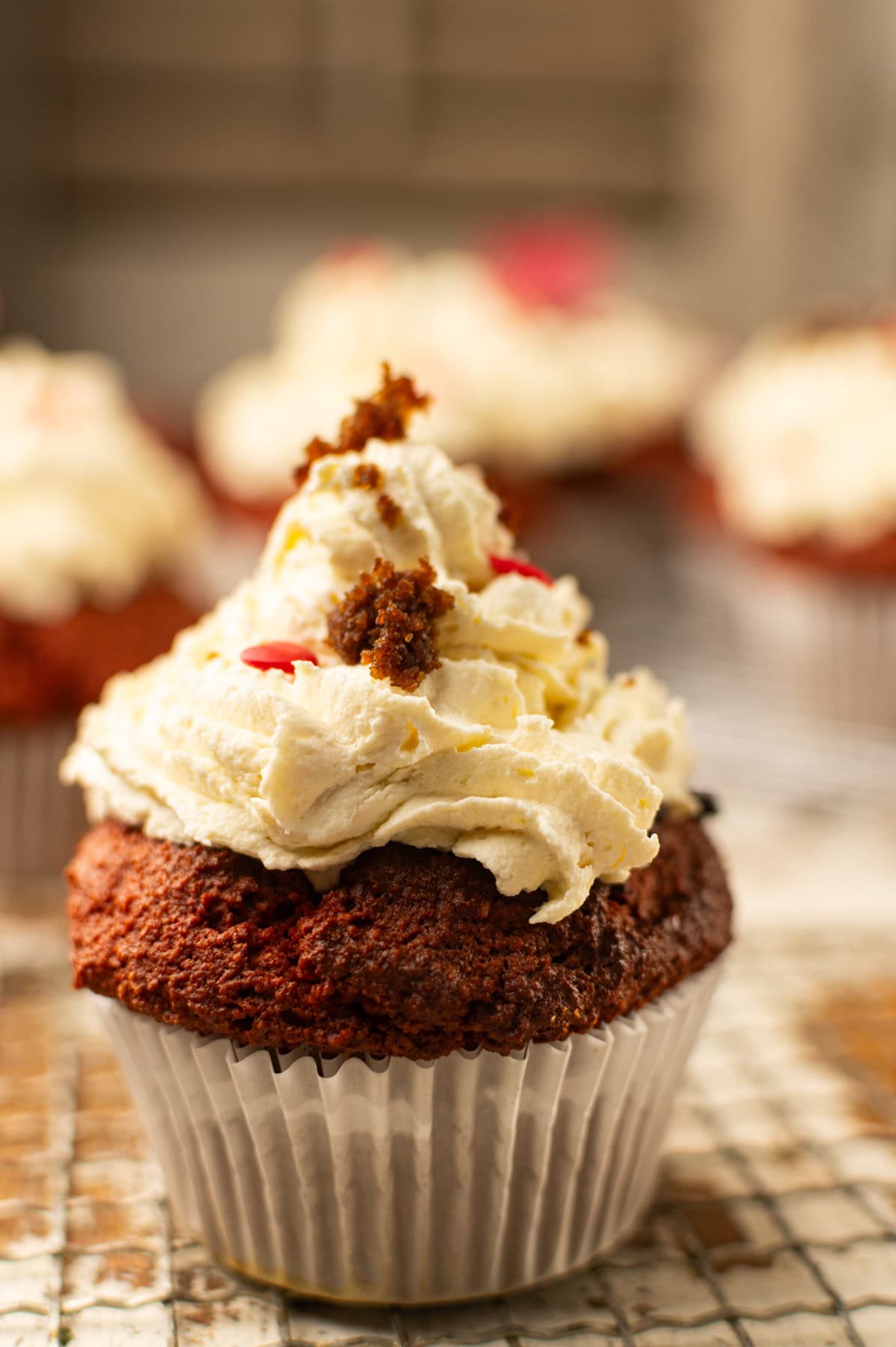 A close-up of a red velvet cupcake with creamy swirled frosting, decorated with small crumbs and red sprinkles, sitting on a wire cooling rack with more cupcakes blurred in the background.
