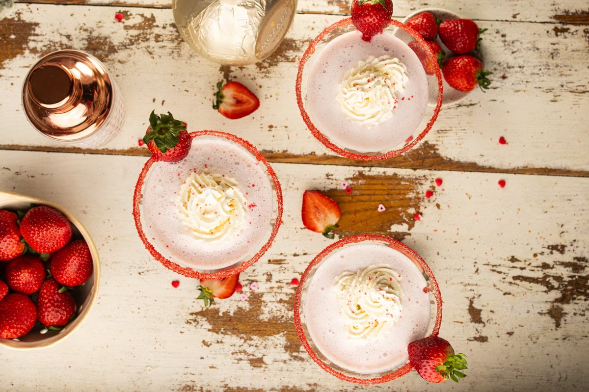 Three strawberry milkshakes with whipped cream, garnished with whole strawberries, sit on a rustic wooden table. A bowl of fresh strawberries, a cocktail shaker, and scattered strawberry pieces are also visible.