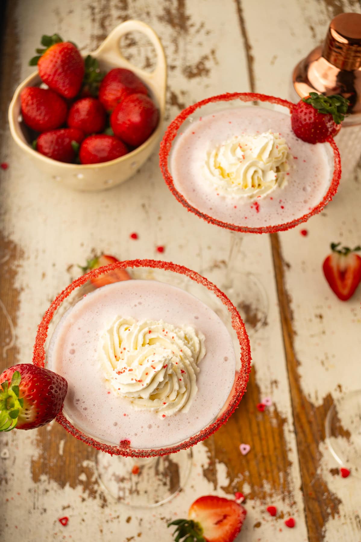 Two pink cocktails in sugared-rim glasses topped with whipped cream and sprinkles, each garnished with a fresh strawberry. A bowl of strawberries and a cocktail shaker sit on a rustic wooden table.